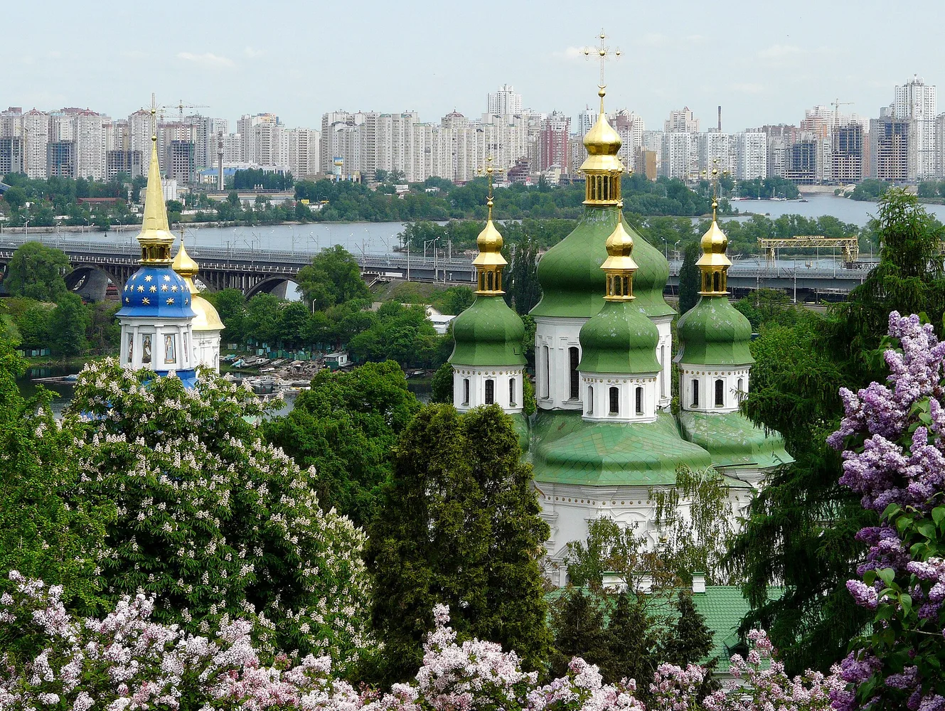 Куполи та дзвіниця православного собору на тлі річки й міського житлового масиву, навколо дерева й квітучі кущі.