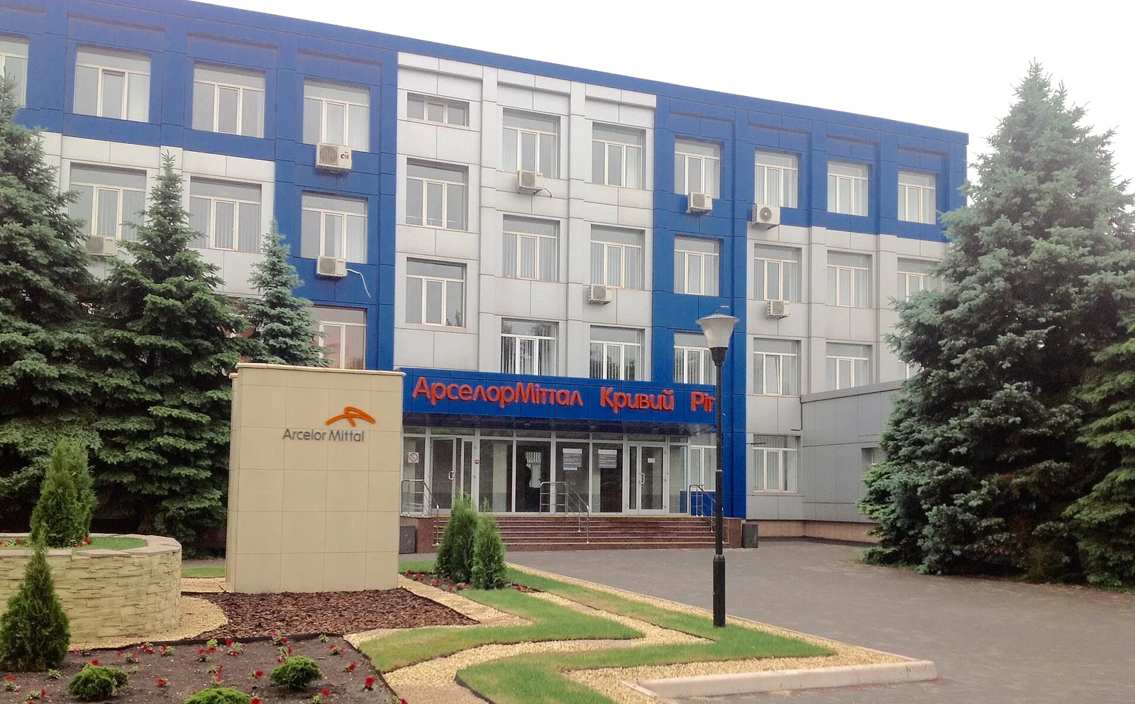Фасад адміністративної будівлі з написом «АрселорМіттал Кривий Ріг», ландшафтне оформлення й ялинки на передньому плані.