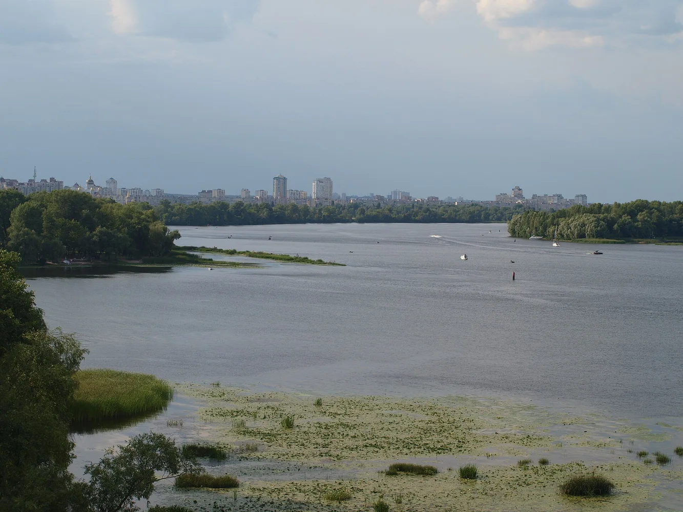 панорамне фото великої річки з берегами, кількома малими човнами та міськими будівлями на горизонті