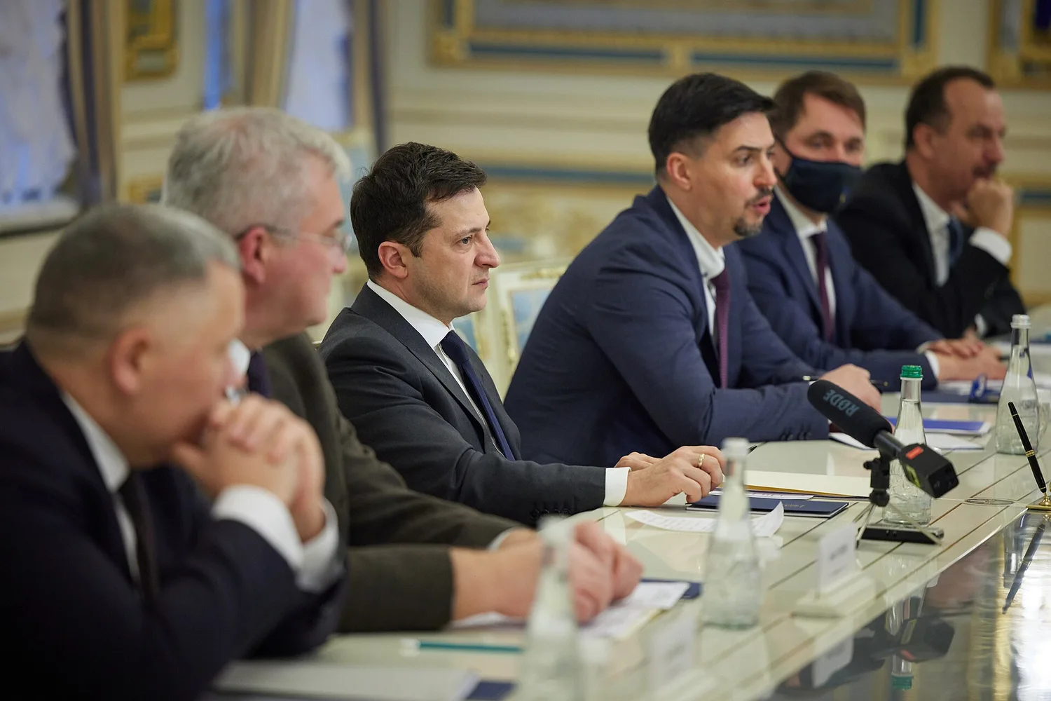 Володимир Зеленський і інші посадовці сидять за столом під час наради; мікрофон і пляшки з водою на столі.