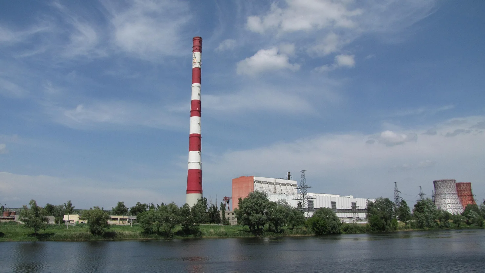 Термальна електростанція біля берега річки: висока смугаста димова труба, корпуси будівель і куполоподібні градирні на задньому плані.