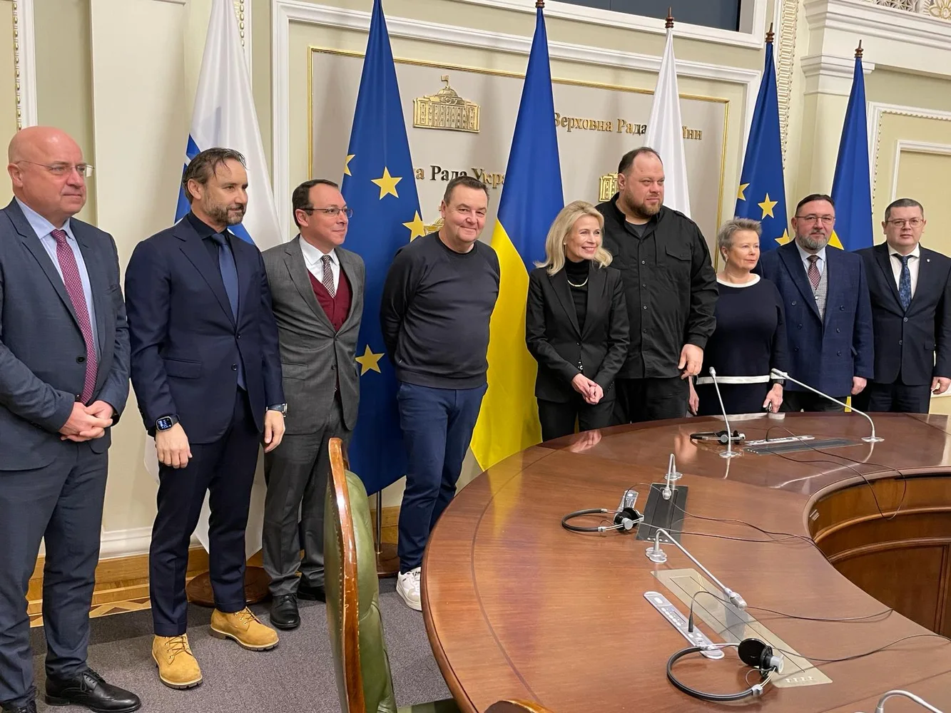 Група людей стоїть у залі біля прапорів України та ЄС, на стіні позаду видно напис «Верховна Рада України»; круглий стіл і мікрофони попереду.