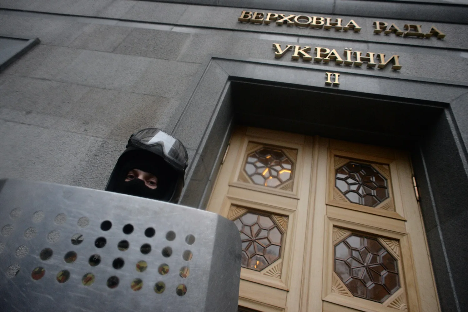 Вхід до будівлі Верховної Ради України з частиною дверей, написом «Верховна Рада України» і охоронцем із щитом на передньому плані.