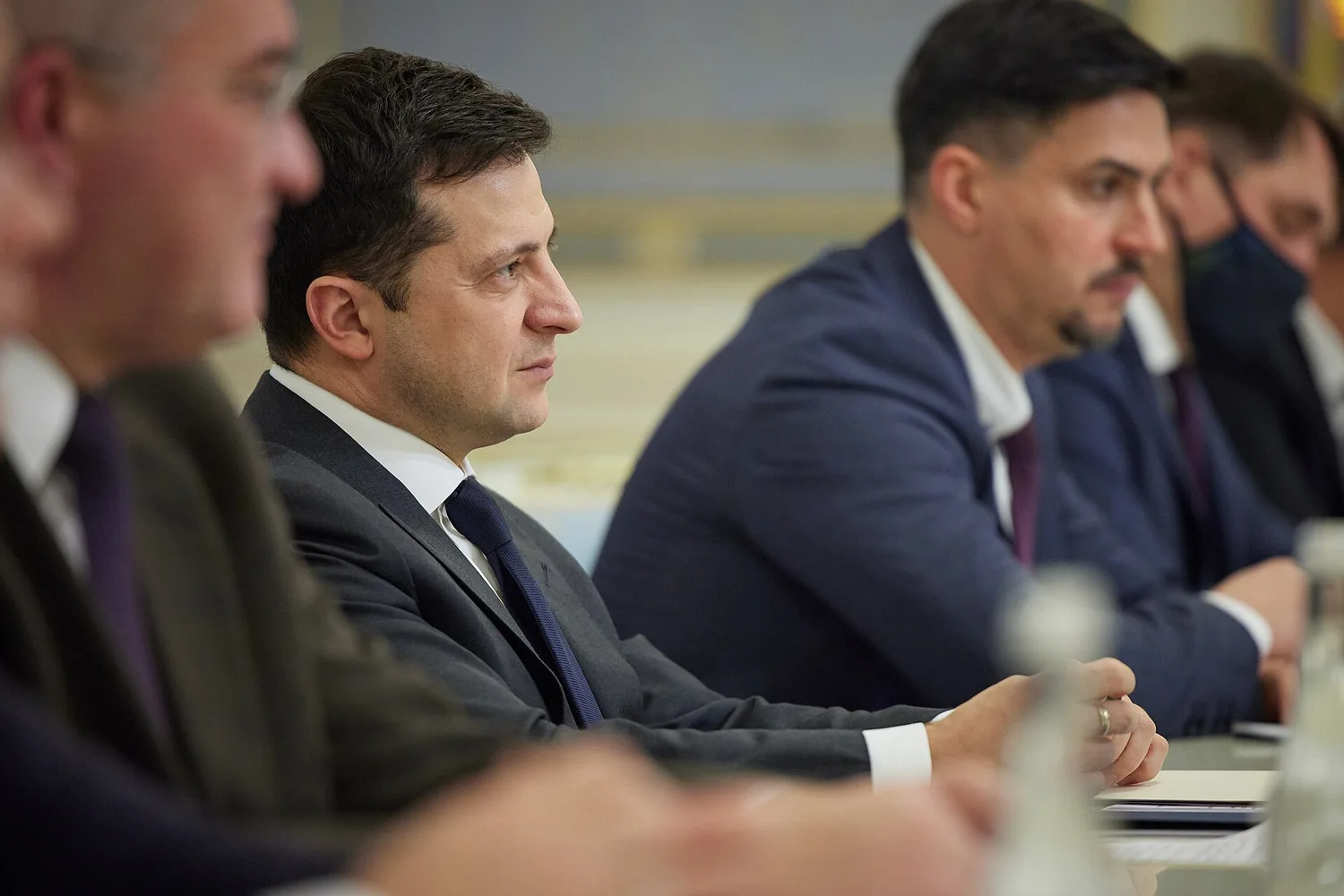 Чоловік у костюмі і краватці сидить за столом під час офіційної зустрічі; поруч інші учасники за столом у конференц-залі.