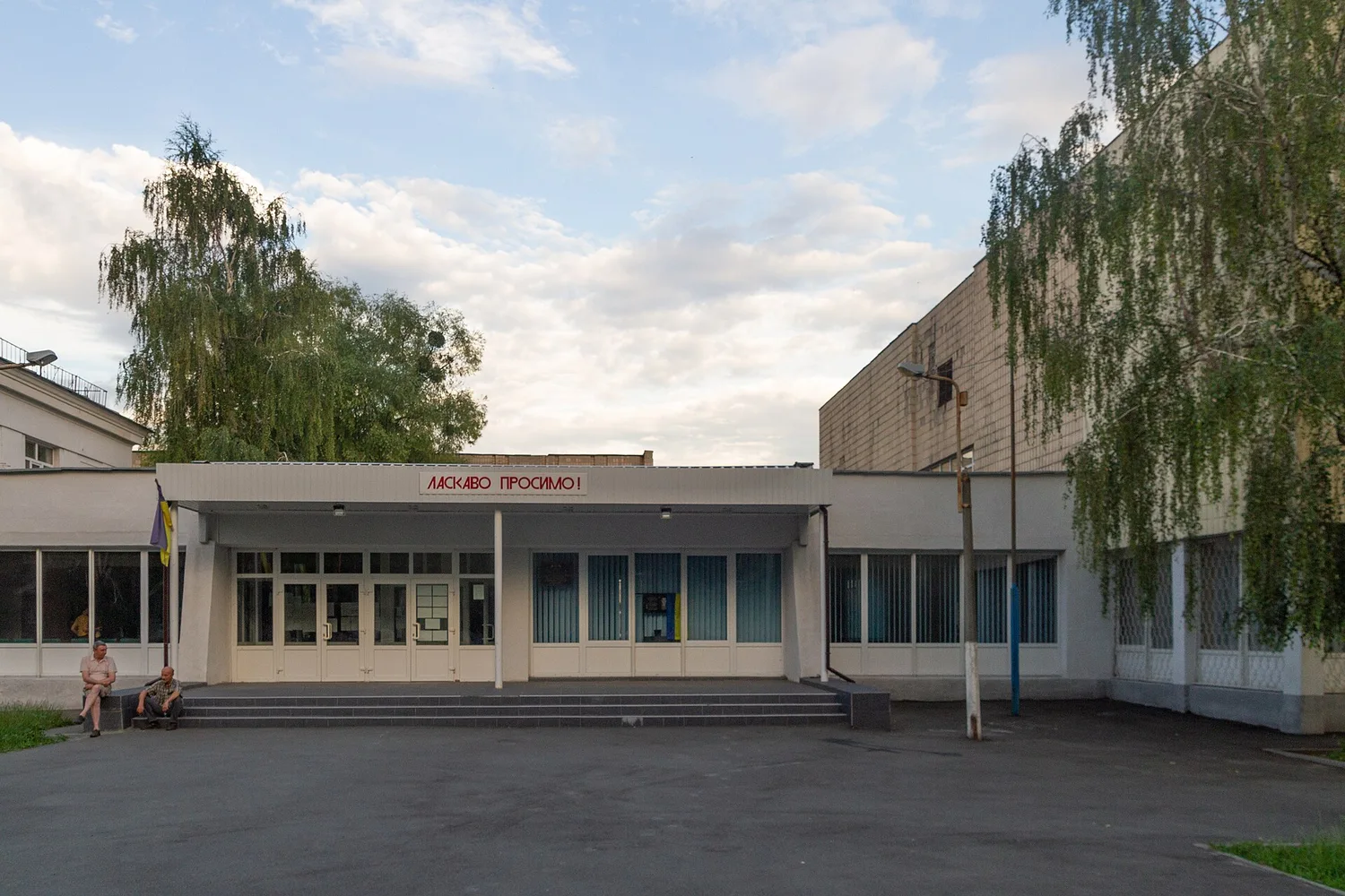 Фасад київської школи з головним входом, ґанком і сходами; на передньому плані асфальтований майданчик, ліворуч сидять дві людини, навколо дерева.