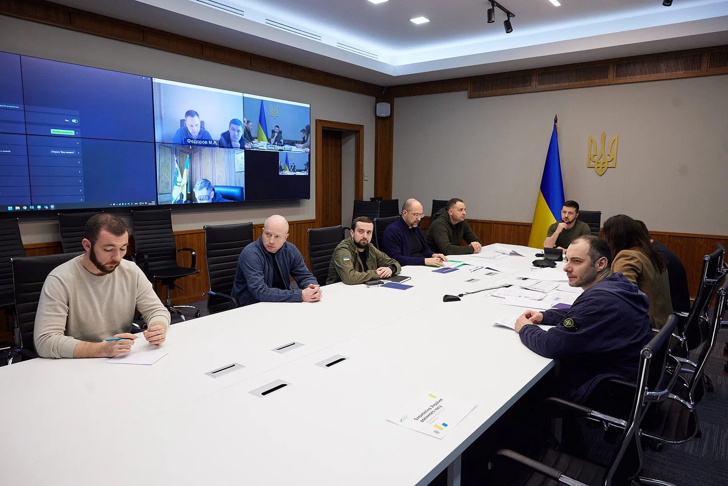 Керується засідання урядової наради в кабінеті: кілька людей за великим білим столом, на стіні прапор України і тризуб, великий екран з відеоконференцією.