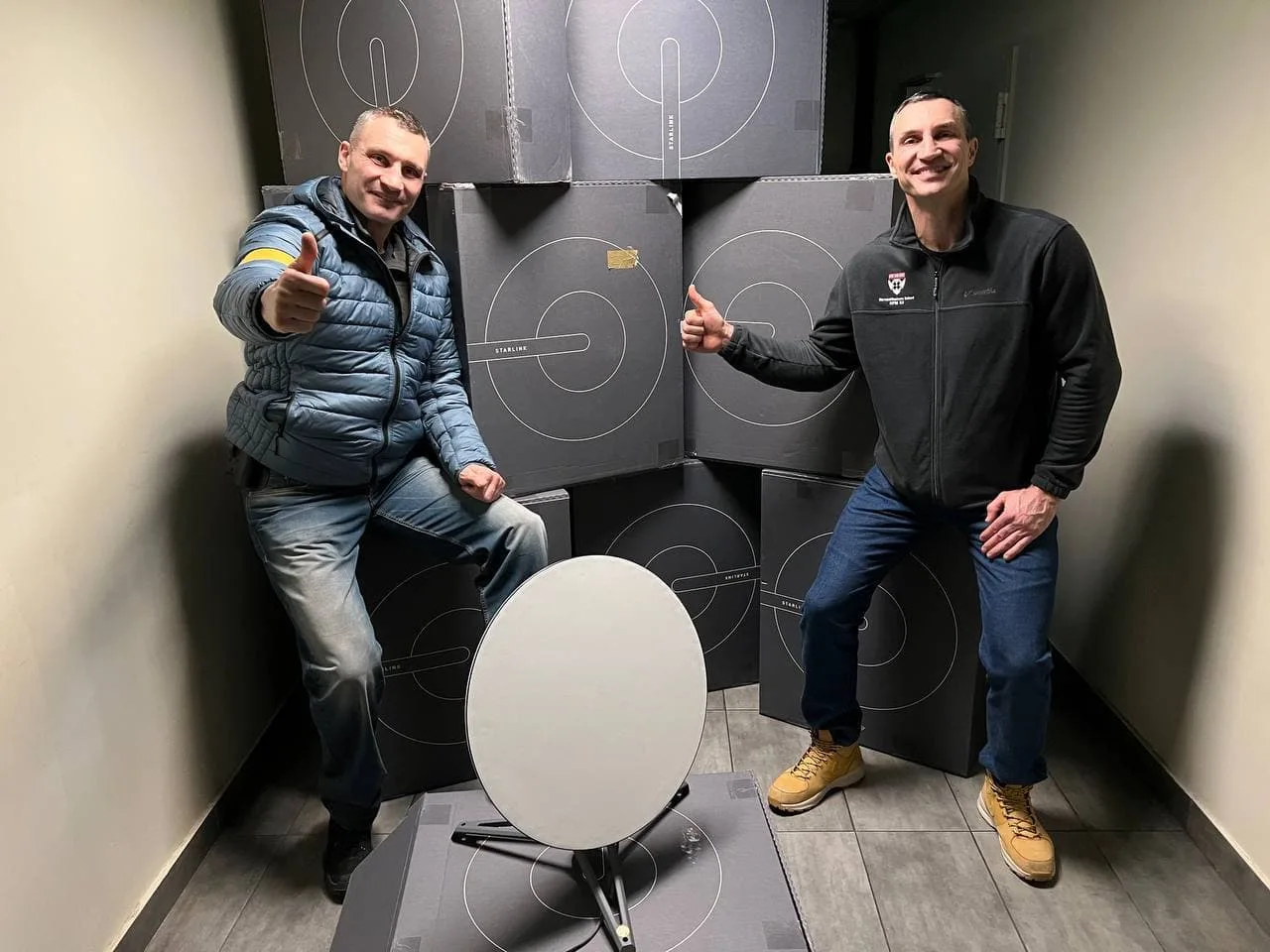 Двоє чоловіків позують поруч зі стопкою коробок Starlink та однією білою тарілчастою антеною на підлозі.