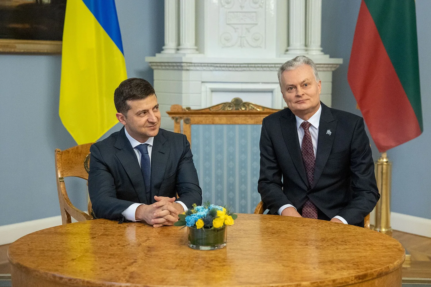 Двоє чоловіків у костюмах сидять за круглим дерев’яним столом у офіційній залі; зліва видний синьо-жовтий прапор, праворуч — зелено-червоний прапор, на столі — невелика ваза з жовто-блакитними квітами.