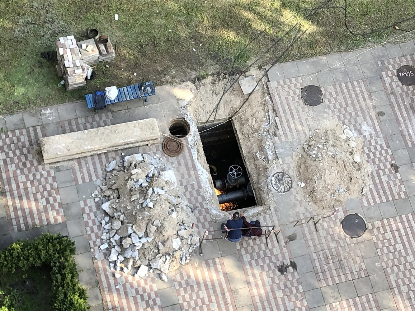 Працівник проводить зварювальні роботи на підземному трубопроводі теплопостачання; вигляд згори на відкритий люк і труби, поруч будівельні матеріали.