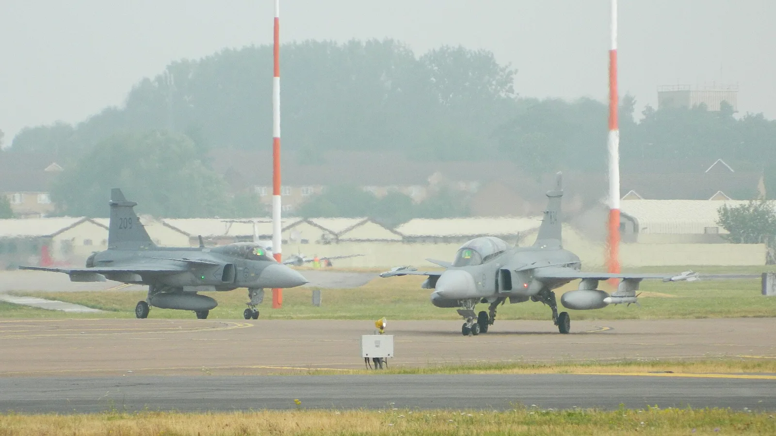Два багатоцільові винищувачі Saab JAS 39 Gripen на стоянці аеродрому, вид збоку, туманна погода.