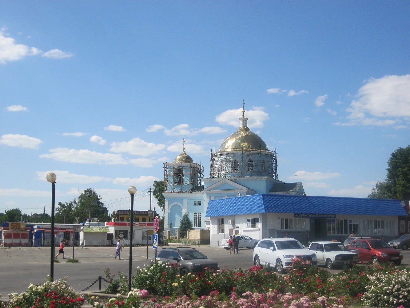 Площа з клумбами, дорожнім покриттям, припаркованими автомобілями; на задньому плані—церква з золотими куполами під блакитним небом.