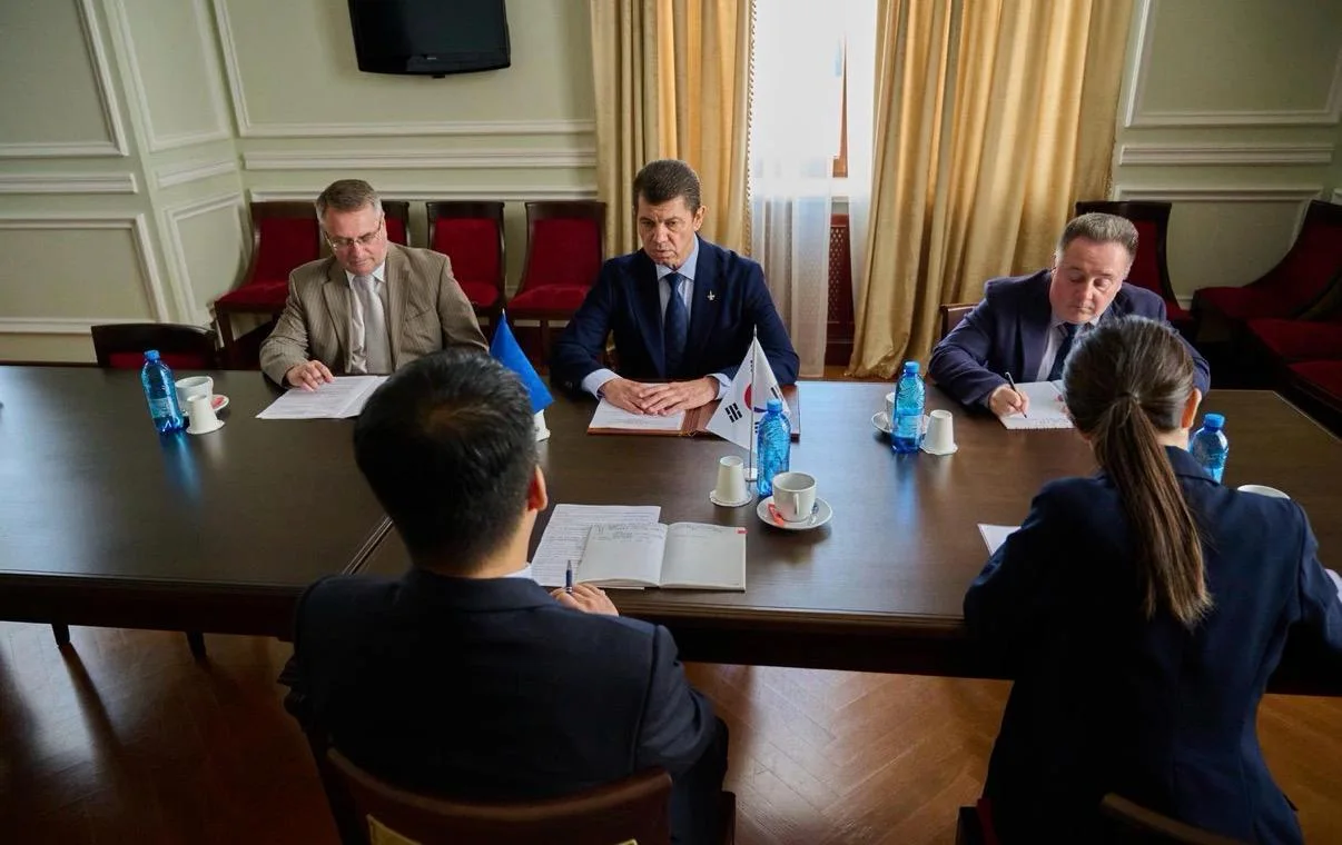 Переговори за столом: кілька посадовців (троє з одного боку, двоє з іншого) у формальному кабінеті, на столі папери, чашки, пляшки води та маленькі прапорці Південної Кореї й України.