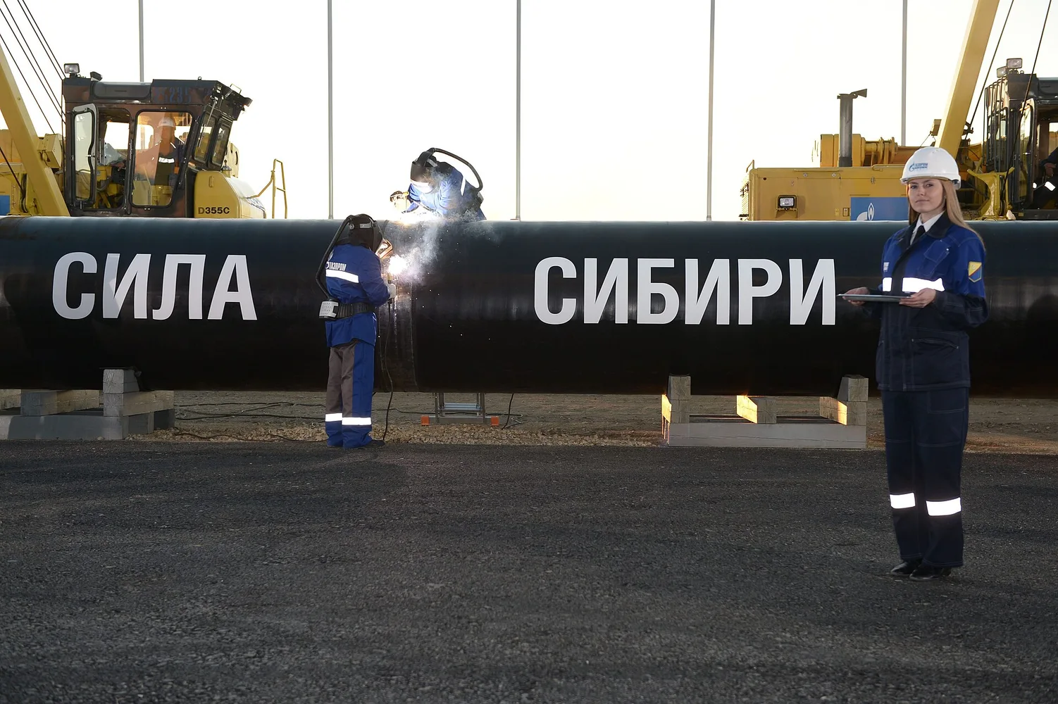 Робітники зварюють ділянку газопроводу «Сила Сибіри», на трубі видимий напис «СИЛА СИБІРИ», поруч стоїть працівниця у захисному одязі.