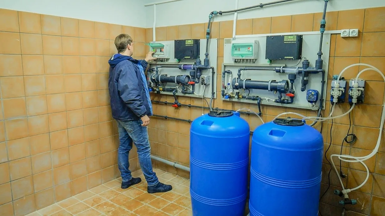 Чоловік у робочому одязі стоїть у приміщенні біля обладнання для водоочистки/фільтрації та двох великих синіх пластикових бочок.